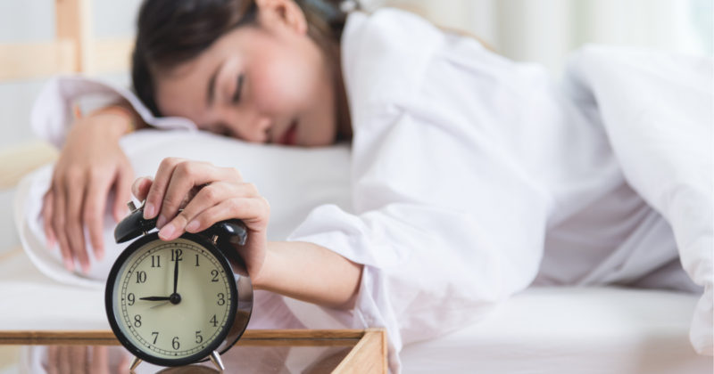 Cansaço excessivo: quando investigar com exames de sangue? Mulher com cansaço excessivo tenta acordar com despertador ao lado, ilustrando fadiga persistente.