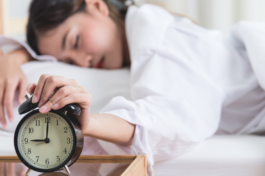 Mulher com cansaço excessivo tenta acordar com despertador ao lado, ilustrando fadiga persistente.